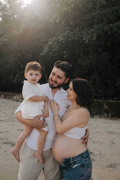 Foto do casal com o filho sorrindo na beira do mar.'