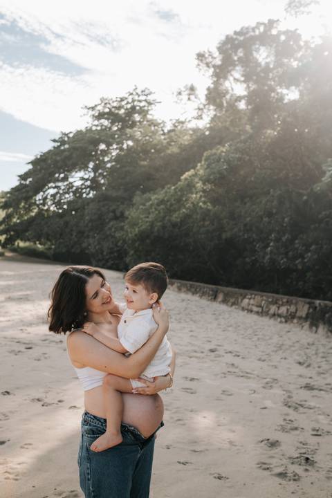 Foto da gestante sorrindo com o filho na areia da praia.'