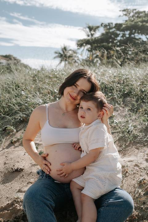 Foto da gestante abraçada com o filho na duna de areia.'