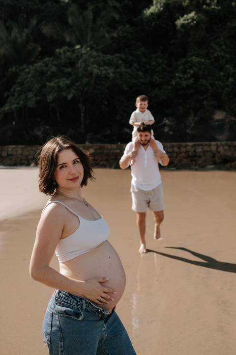 Foto da gestante em destaque com o pai e o filho ao fundo na areia.'
