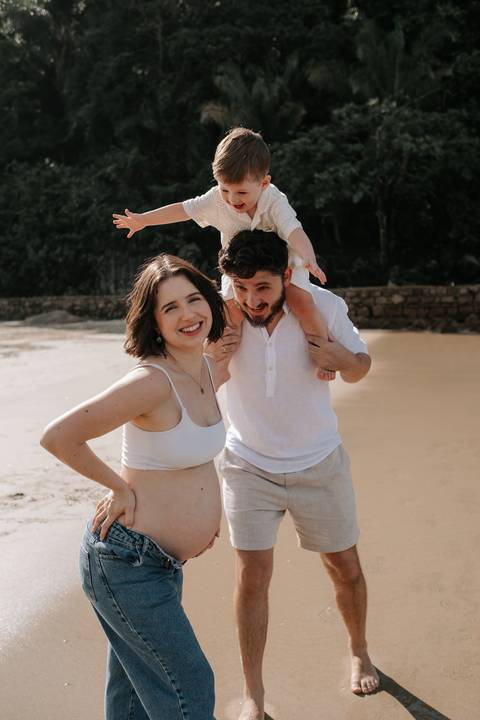 Foto do pai com o filho nos ombros e a gestante sorrindo na praia.'