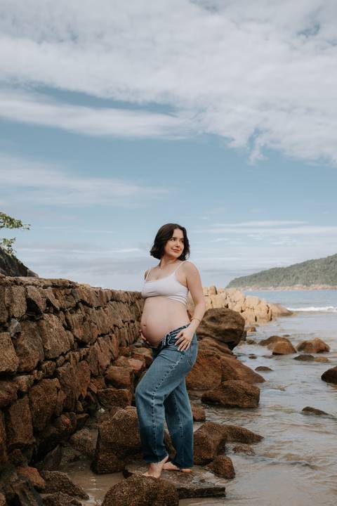 Foto da gestante de perfil nas rochas da praia.'