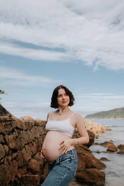 Foto da gestante em pé observando o mar em ensaio externo.'