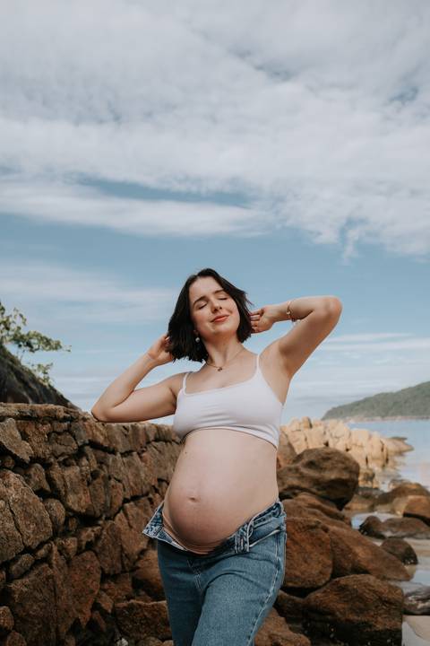 Foto da gestante sorrindo nas rochas durante ensaio na praia.'