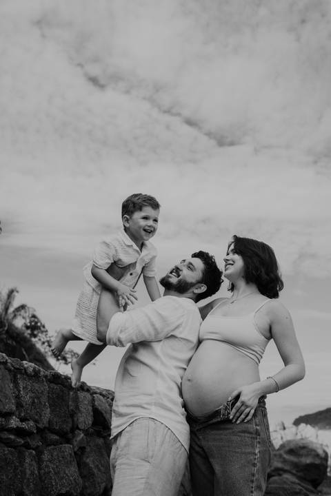 Foto em preto e branco do pai levantando o filho com a gestante ao lado.'