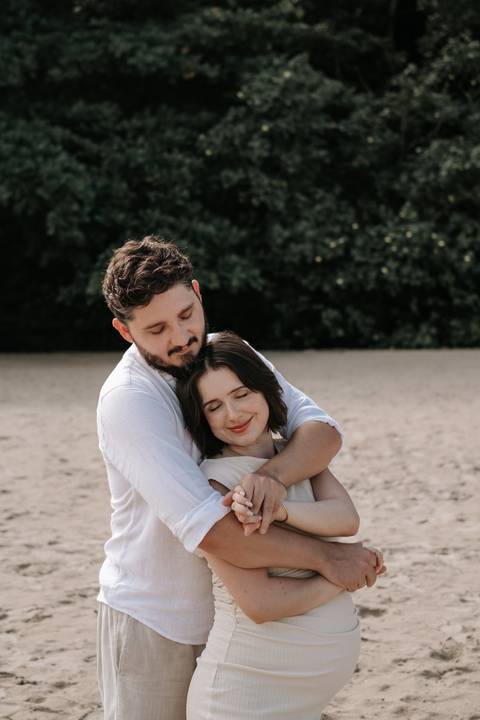 Foto do casal abraçado na areia da praia com céu claro.'
