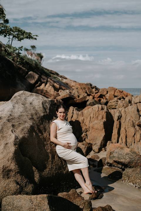 Foto da gestante posando nas rochas da praia com luz natural.'