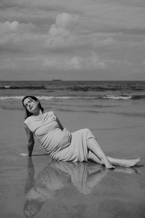 Foto em preto e branco da gestante sentada na areia com o mar de fundo. '