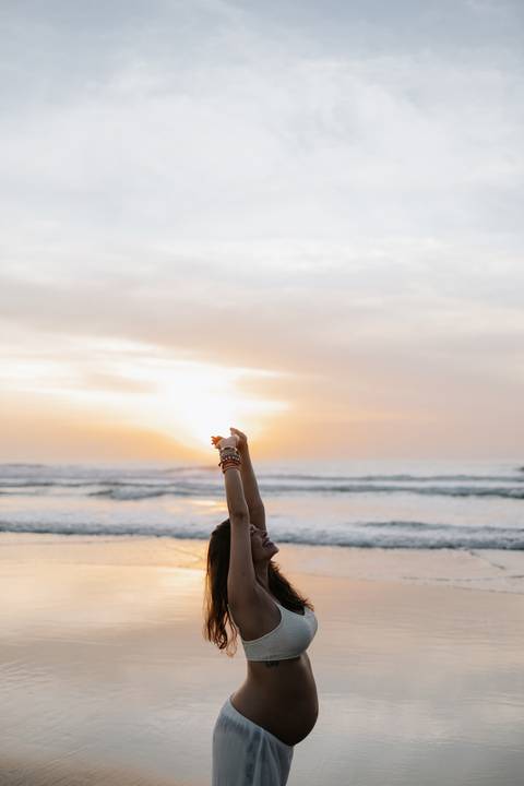 Foto do detalhe das mãos da gestante em direção ao sol no amanhecer.'