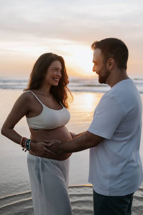 Foto do casal sorrindo durante ensaio gestante no nascer do sol.'