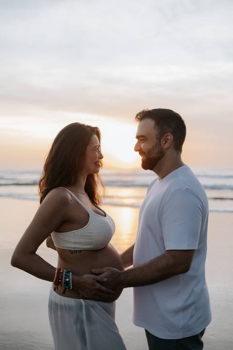 Foto do casal trocando olhares com o mar ao fundo no amanhecer.'