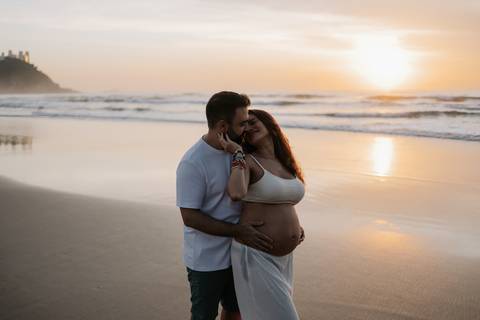 Foto do pai abraçando a gestante por trás na beira do mar.'