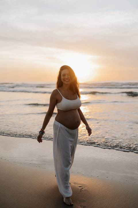 Foto da gestante caminhando na areia durante o amanhecer no litoral.'