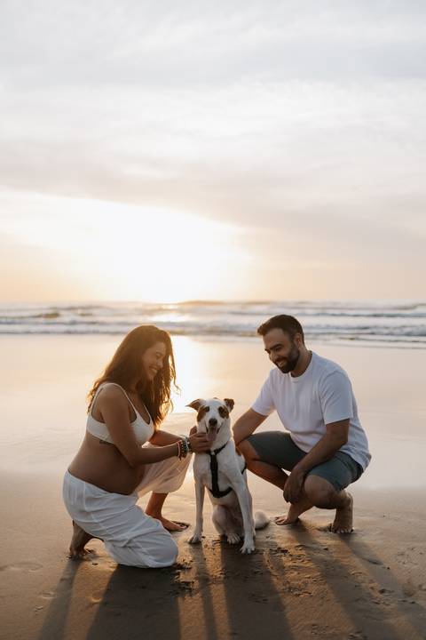 Foto do casal brincando com o cachorro na areia da praia.'