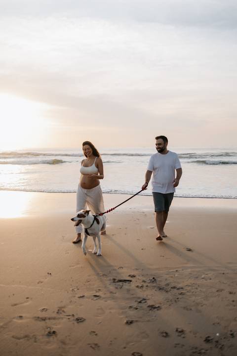 Foto do casal caminhando com o cachorro na guia pela beira do mar.'