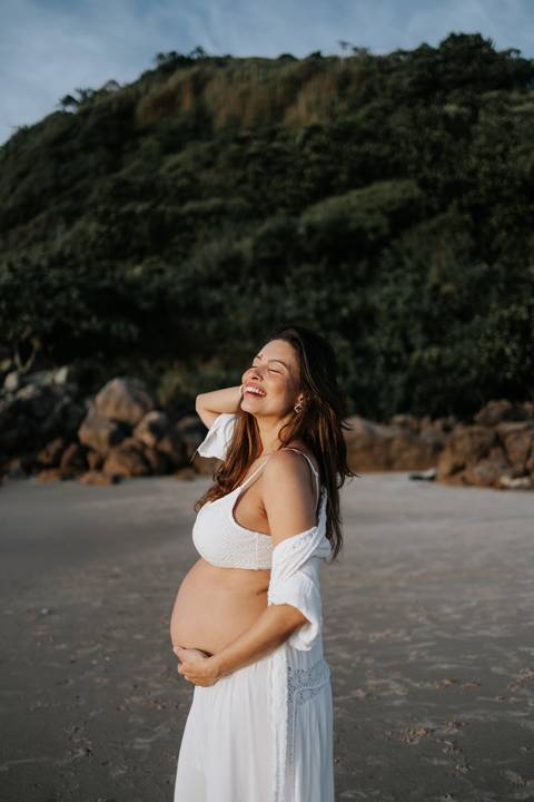 Foto da gestante sorrindo em meio à natureza da praia.'