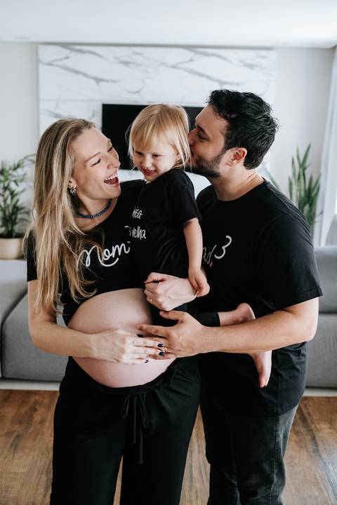 Foto do casal sorrindo com a filha no colo e destaque para a barriga.'