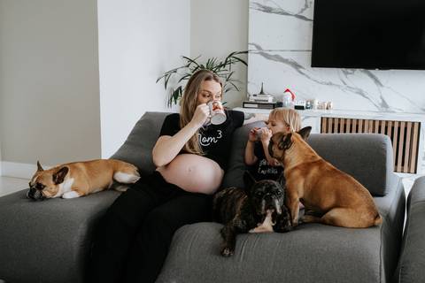Foto da gestante sentada no sofá com a filha mais velha e os três cachorros.'