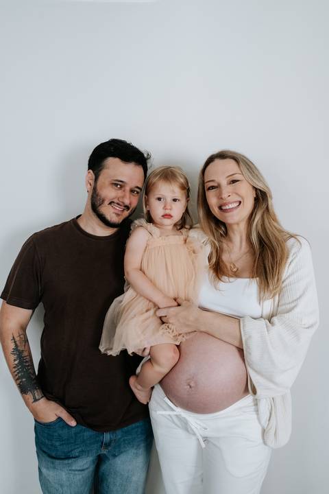 Foto do casal sorrindo com a filha no colo no ensaio em casa.'
