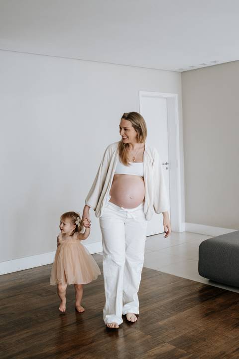 Foto da gestante caminhando de mãos dadas com a filha na sala.'