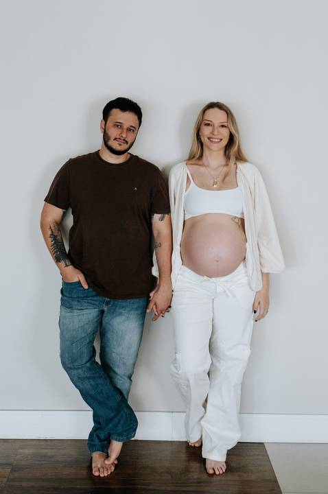 Foto do casal em pé de mãos dadas destacando a barriga de gêmeos.'