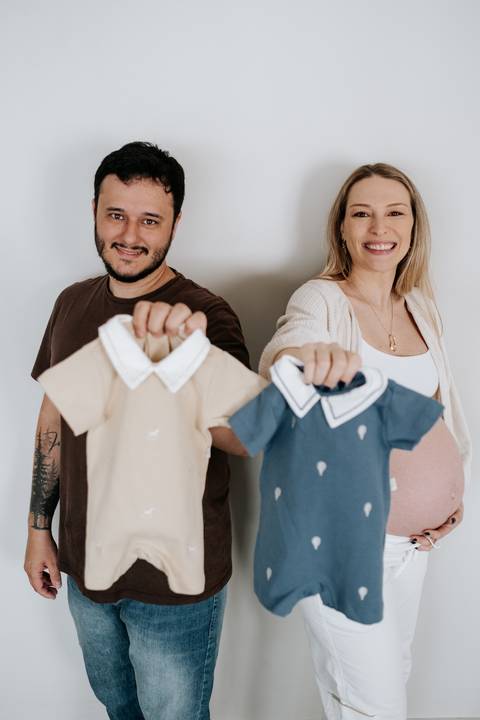 Foto do casal segurando as duas roupinhas para os bebês gêmeos.'
