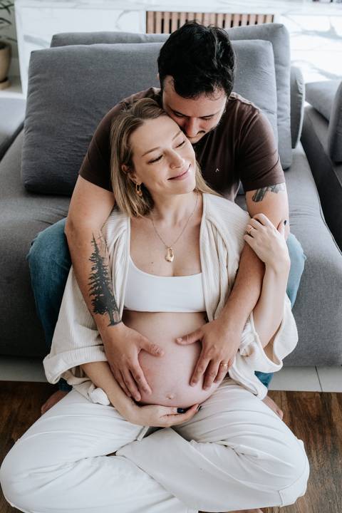 Foto do pai abraçando a gestante por trás sentados no sofá.'