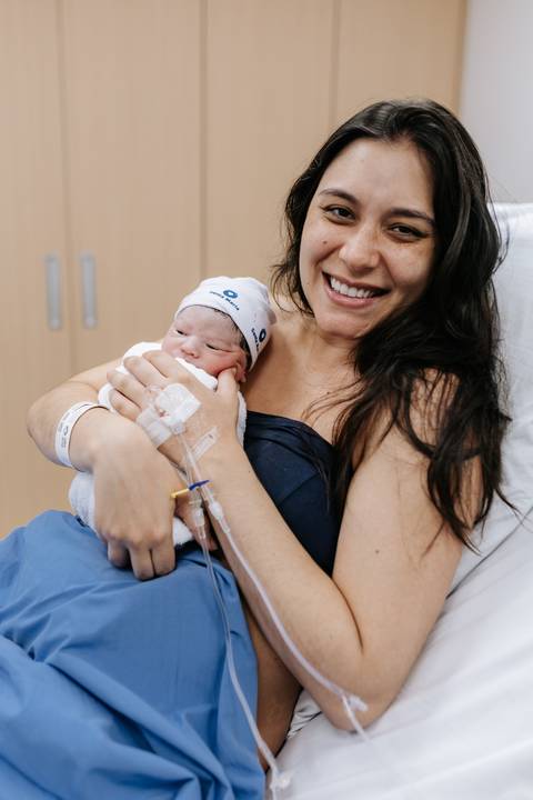 Foto da mãe sorrindo e abraçando o recém-nascido no hospital.'