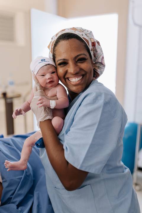 Foto da enfermeira sorridente segurando o bebê ao lado da mãe.'