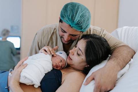 Foto do pai abraçando a mãe com o bebê no colo na cama hospitalar.'