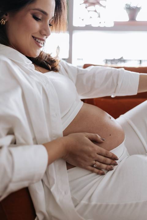 Foto em detalhe das mãos da gestante sobre a barriga em ensaio em casa.'