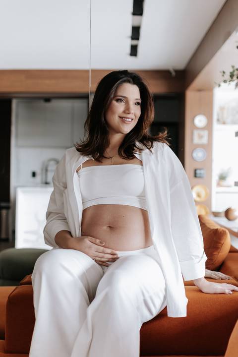 Foto da gestante sorrindo com blusa branca aberta destacando a barriga.'