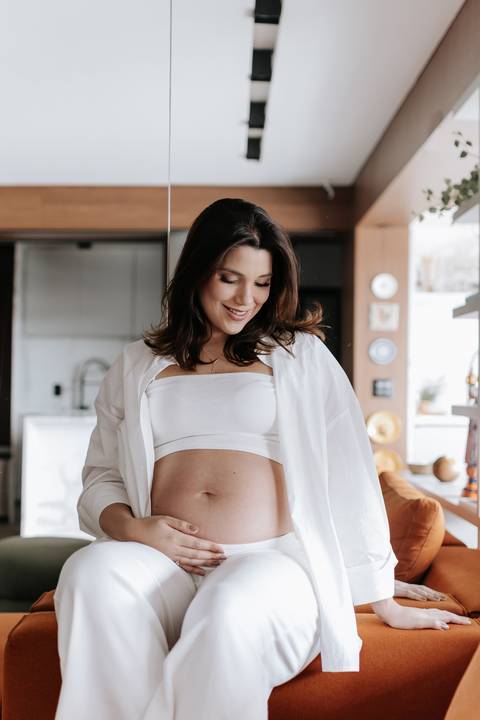 Foto aproximada da gestante olhando para a barriga no ambiente da sala.'