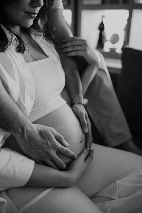 Foto em preto e branco do detalhe das mãos do casal na barriga.'
