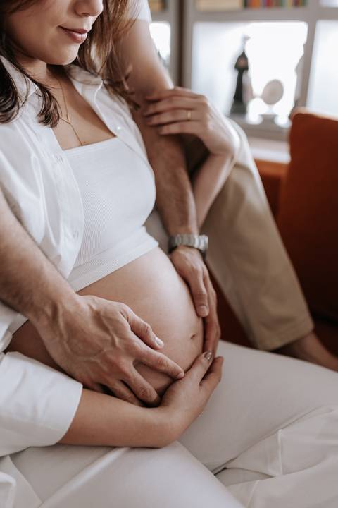 Foto em detalhe das mãos do casal sobre a barriga da gestante.'