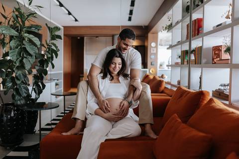 Foto do casal sorrindo no sofá em ensaio gestante documental.'