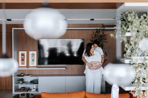Foto do casal abraçado na sala de estar decorada da residência.'