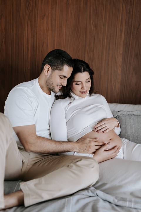 Foto do casal sentado no sofá sorrindo e olhando para a barriga.'