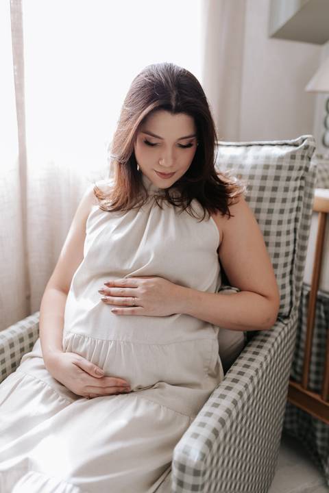Foto da gestante sentada em poltrona de amamentação no quarto do bebê.'