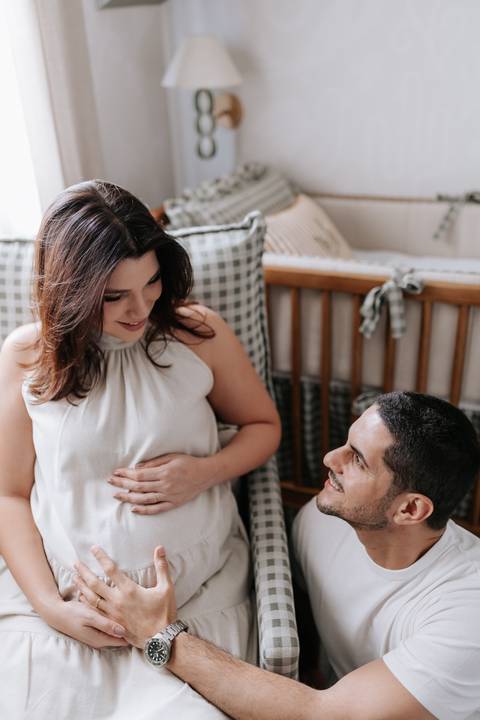 Foto do casal sorrindo e interagindo no quarto preparado para o bebê.'