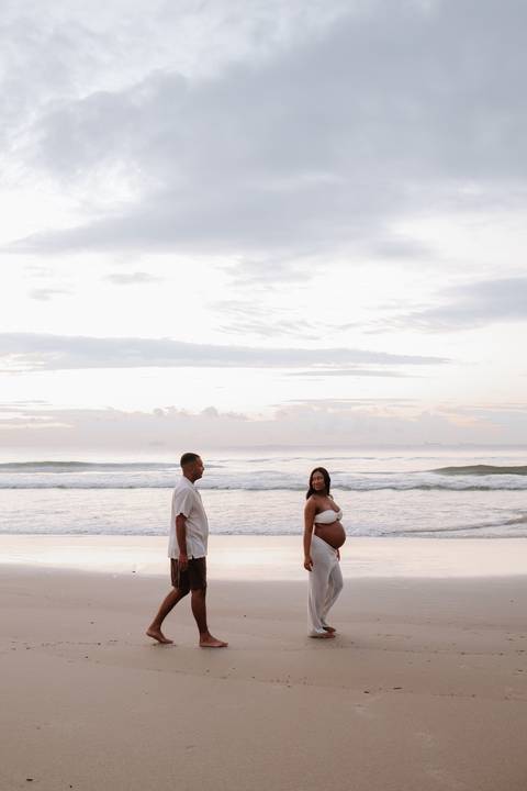Foto do casal em pé na beira do mar com céu de cores suaves.'