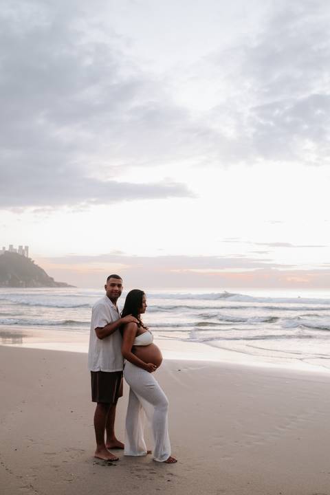 Foto do pai abraçando a gestante por trás com vista para o mar.'