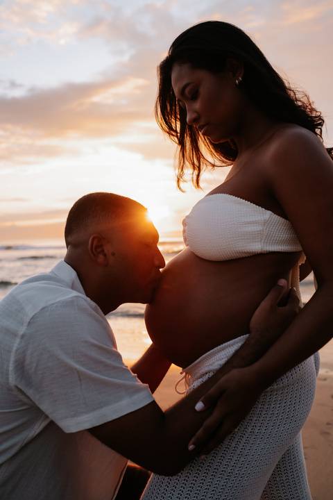 Foto do pai beijando a barriga da gestante durante o amanhecer.'