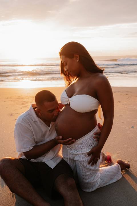 Foto do marido beijando a barriga da gestante com o sol brilhando ao fundo.'