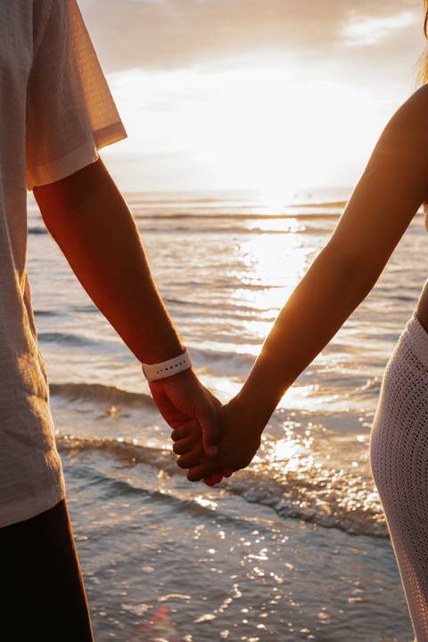 Foto em detalhe das mãos do casal dadas com o reflexo do sol na água.'