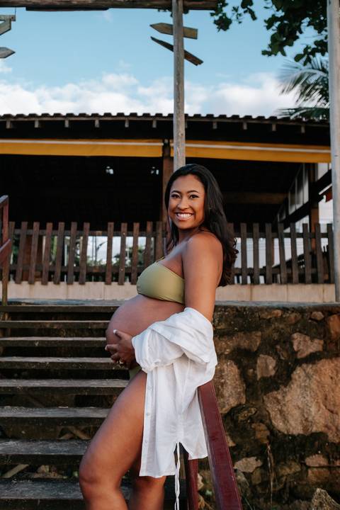 Foto de perfil da gestante sorrindo em frente a uma escadaria de madeira.'
