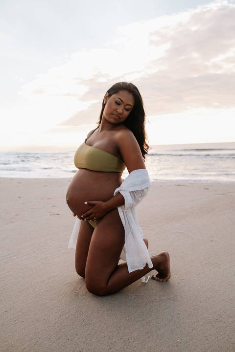 Foto da gestante de perfil na areia destacando a barriga de grávida.'