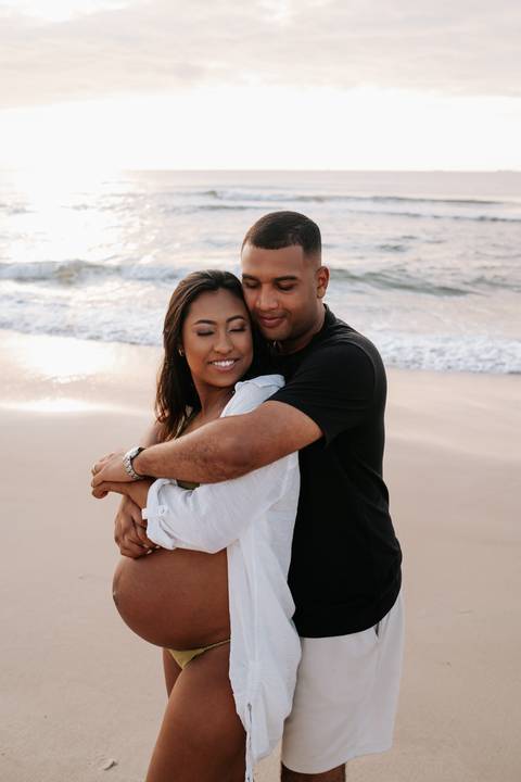 Foto do pai abraçando a gestante por trás na beira do mar.'