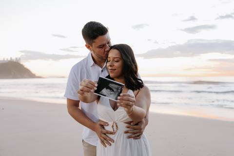 Foto do casal segurando a imagem do ultrassom na beira do mar.'
