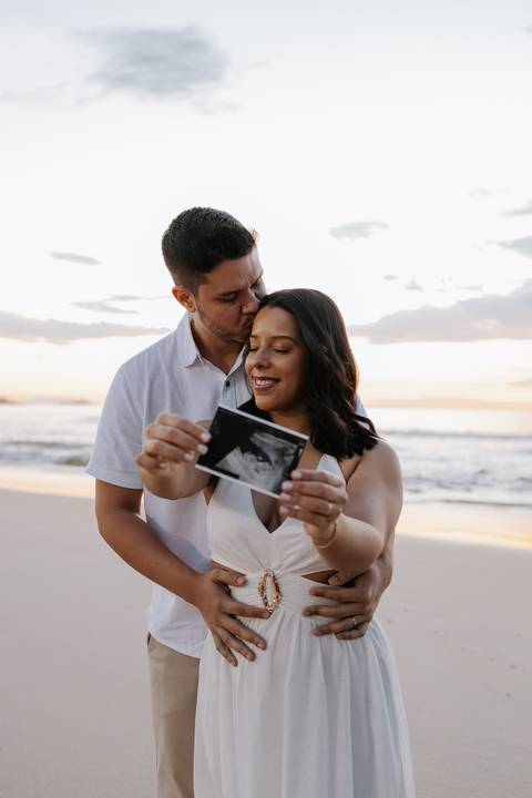 Foto aproximada do casal exibindo o ultrassom do bebê no nascer do sol.'
